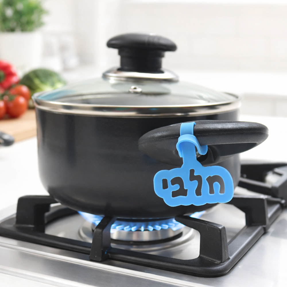 
סיר בישול שחור עם מכסה זכוכית על כיריים גז, עם תג סיליקון כחול עם הכיתוב "חלבי", על רקע מטבח מואר עם קרש חיתוך, ירקות וסכין